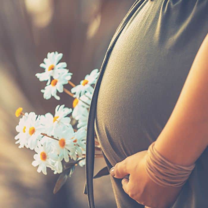 A very pregnant woman is holding her belly and a bouquet of daisy flowers.