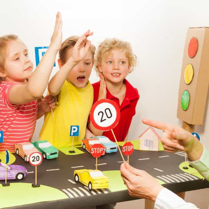 Three young children enthusiastically participate in a road safety education lesson, complete with tabletop traffic signs, vehicles, and homemade traffic lights.