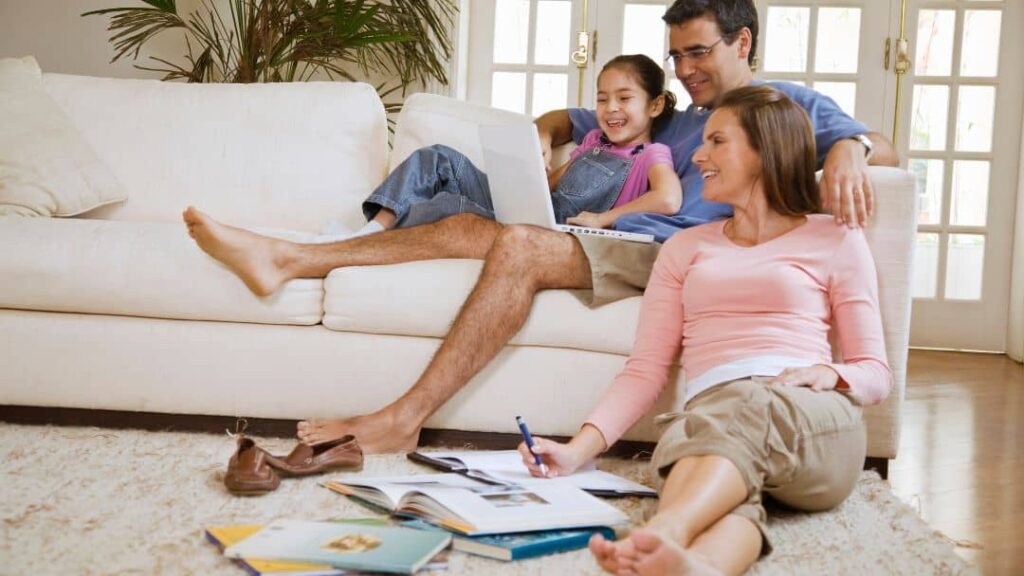 A family of three relaxes in a bright living room. A man and young girl sit on a white couch with a laptop, while a woman sits on the floor, writing, surrounded by open books and papers.