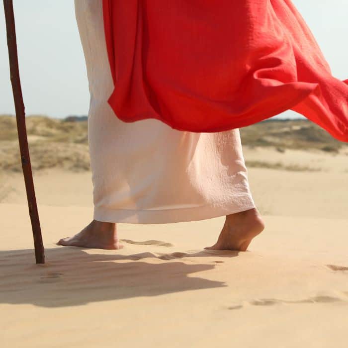 Jesus in a red robe walking on a sandy beach.