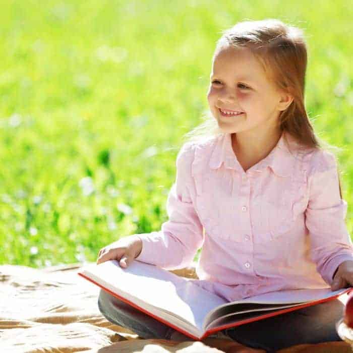 A little girl with a Bible open in her lap. She is reading and enjoying the sunshine on a picnic blanket.
