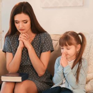 A mother and her daughter are sitting on the couch with hands folded in prayer and a Bible on their lap.
