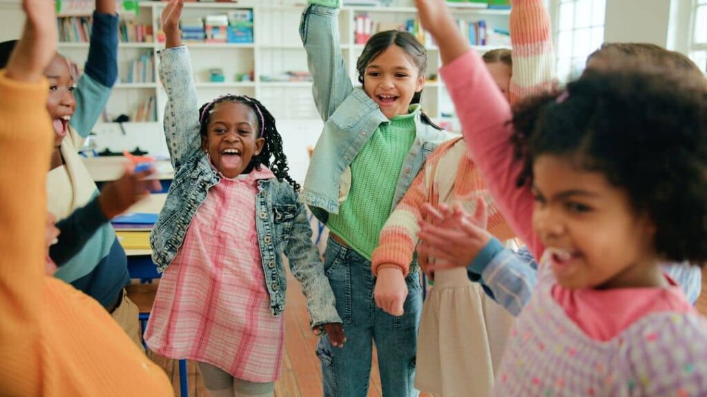 A group of young children joyfully raising their arms and smiling together in a brightly lit classroom with bookshelves—an inspiring scene for those seeking LDS activity day ideas for girls.