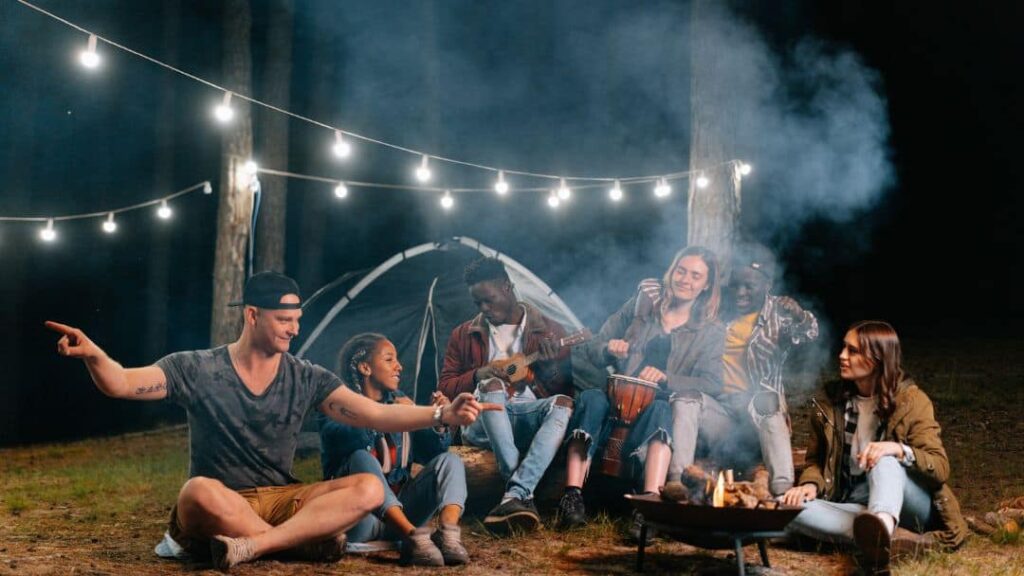 A group of friends sit around a campfire at night near a tent, with string lights overhead—one of the best fun summer ideas with friends. Some play guitar while others talk and laugh, enjoying the outdoors surrounded by trees.