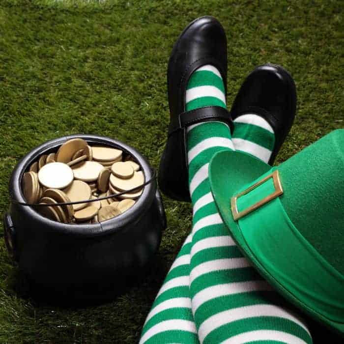 A little girl wearing green and white stripe stockings for St Patrick's Day. There is a pot of gold and a green leprechaun hat next to her legs.
