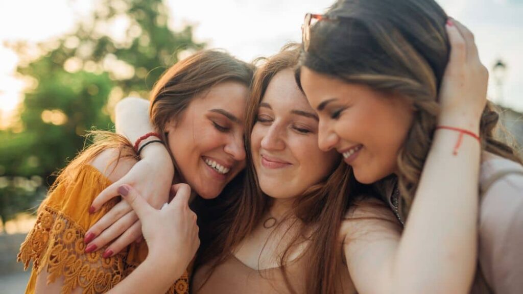 Three young women are standing close together outdoors, smiling and hugging each other warmly, enjoying a happy moment and sharing fun summer ideas with friends. The sun is shining, and trees are visible in the background.
