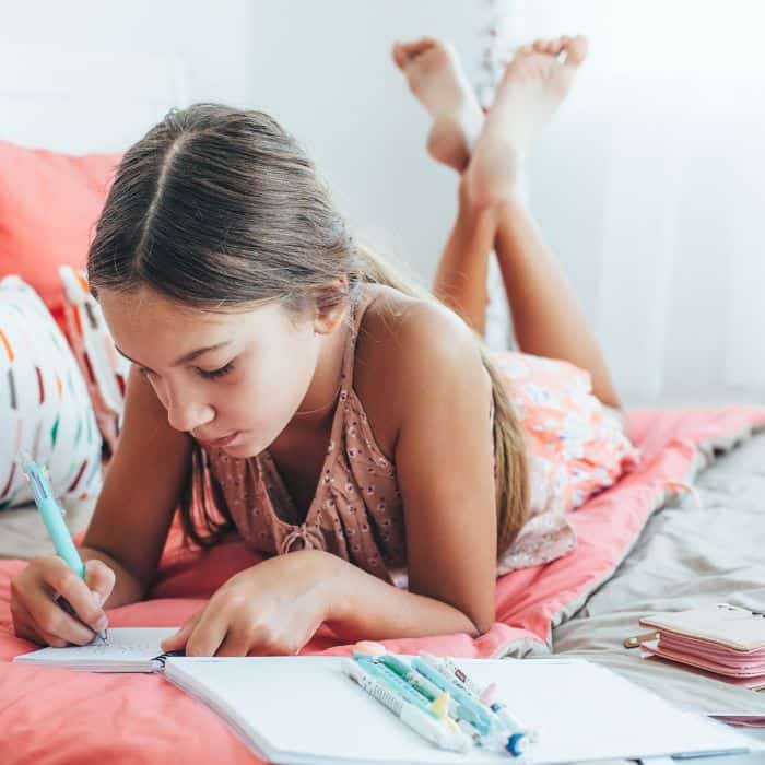 A young girl lying on her stomach on a bed, writing in a notebook with colorful pens scattered around, in a brightly lit room with pink bedding.