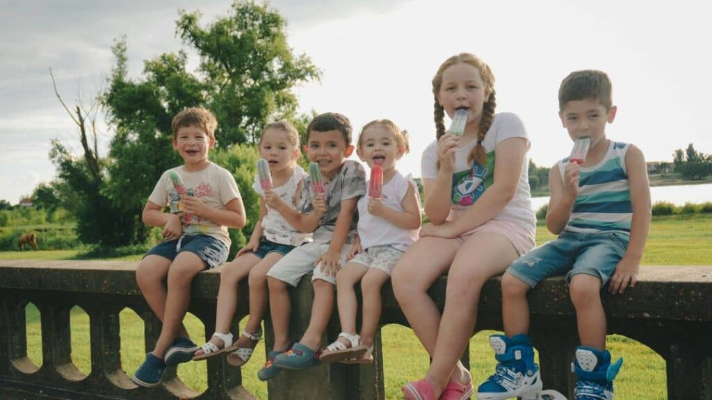 Six young children sit on a low concrete wall outdoors, enjoying colorful ice pops. They are smiling and casually dressed for summer—one of many fun summer ideas with friends—surrounded by green grass and trees on a sunny day.