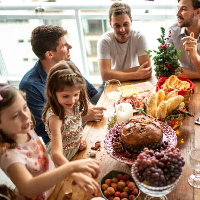An extended family gathered around the table at Christmas time with breads and grapes on the table. Everyone is laughing and smiling as they converse with each other.
