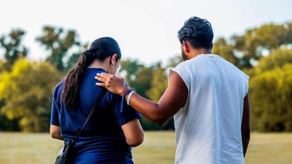 Two people stand outdoors, viewed from behind, sharing a moment surrounded by green grass and trees under a bright sky—capturing the warmth of fun summer ideas with friends.